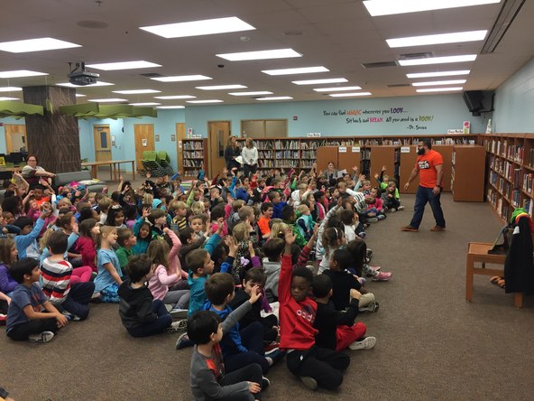 Jimmy Vee performing in a library to a large group of children seated on the floor with hands raised